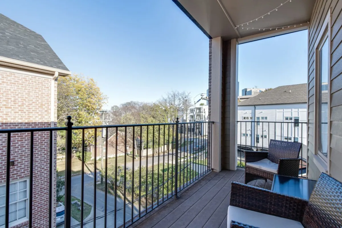 The image showcases the exterior corner balcony of a modern condo with inviting outdoor furniture, looking out over a lively Nashville street. Soft morning sunlight filters through leafy trees, and the setting feels both tranquil and connected to the city. The balcony is perfect for relaxing with coffee, enjoying the breeze, or unwinding as the sun sets, all while being just steps away from the excitement of Broadway and local attractions.