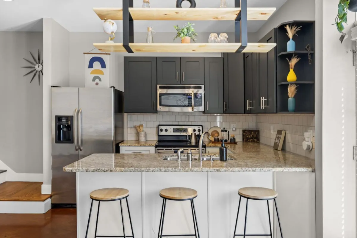 A modern kitchen scene with a granite island, open shelving decorated with plants and dishes, stainless-steel appliances, and comfortable stools. The space is well-lit and stylish, ready for group meals or casual breakfasts.