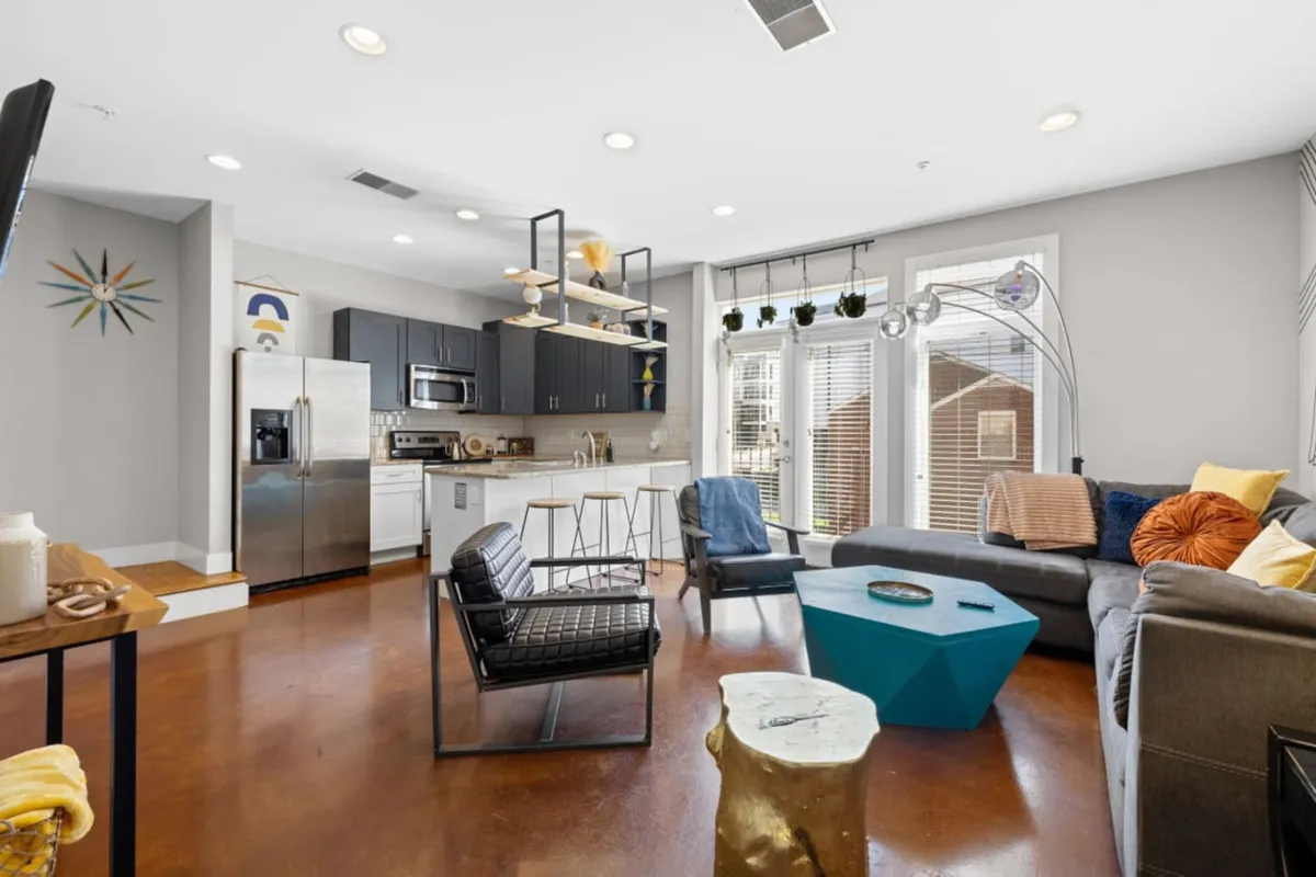 A bright open-plan space featuring a blue coffee table, a sofa with colorful cushions, metal chairs, and a kitchen island. The living and dining areas flow seamlessly together, with natural light highlighting the contemporary decor and inviting seating arrangement.