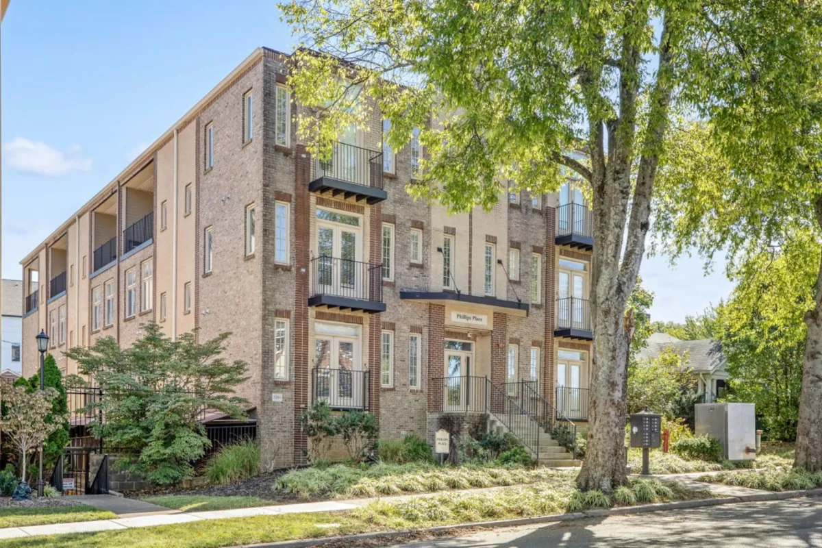 A welcoming brick façade of the condo building featuring balconies, entrance steps, a beautifully landscaped garden, and mature trees. The scene suggests opportunities for social gatherings or quiet outdoor relaxation, and gives a sense of arrival and proximity to the neighborhood.