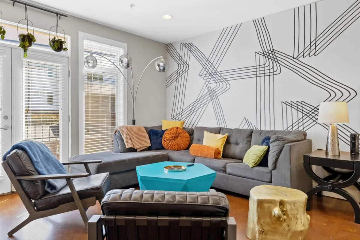 A contemporary and playful living area with geometric wall art, colorful cushions, a unique coffee table, and a stylish floor lamp. The space is filled with natural light and designed for relaxing, socializing, or gathering before or after a night out.