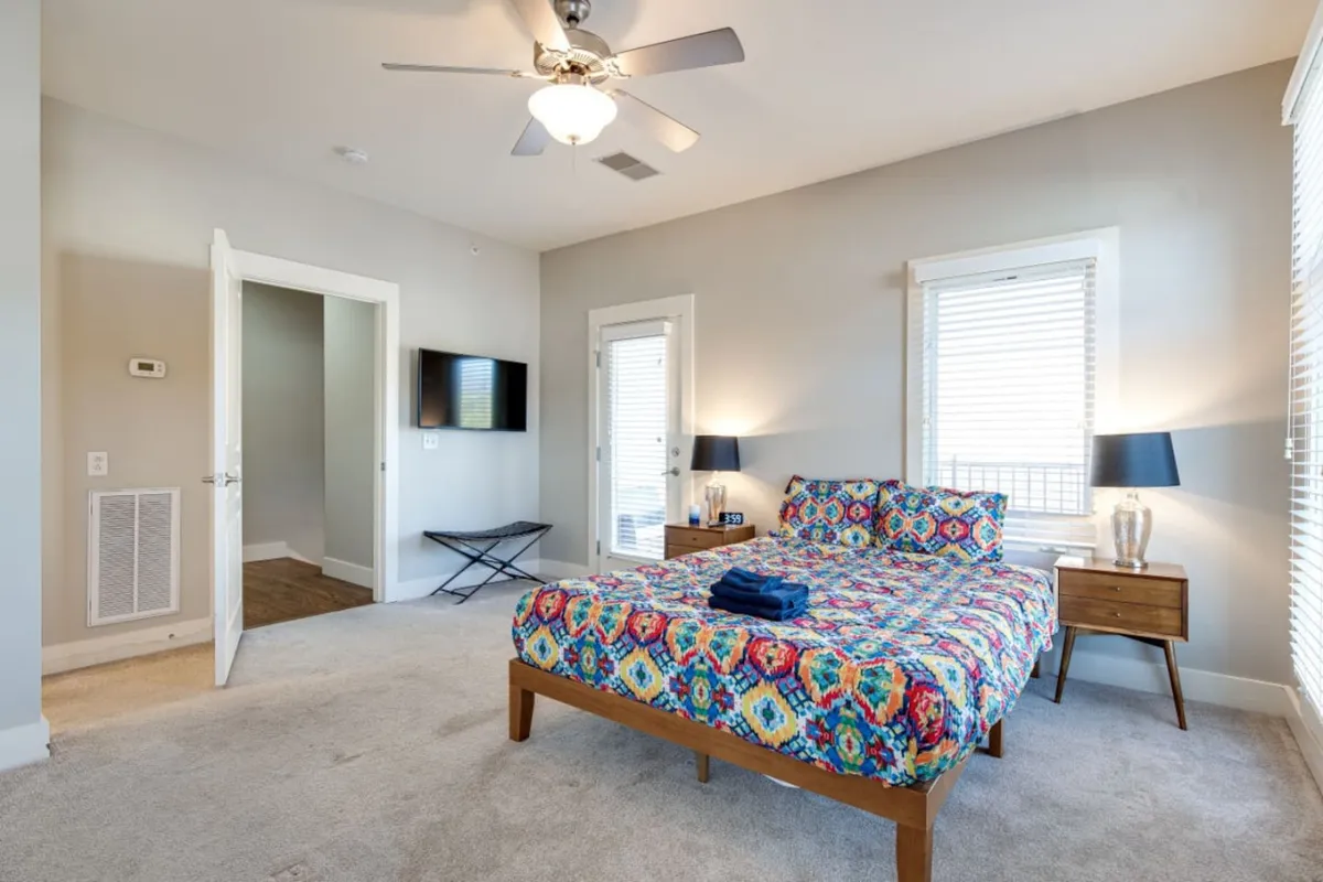 A cozy bedroom featuring a colorful bedspread, nightstand with a lamp, and a wall-mounted television. Natural light pours in through the window, and the room exudes a restful, inviting ambiance. The setup hints at hotel-like comfort and amenities, including the Smart TV mentioned in the section.