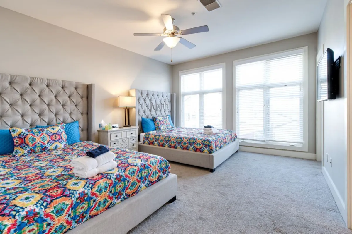 A cheerful bedroom showcasing multiple queen beds with colorful bedspreads, large windows, decorative headboards, and a ceiling fan. The layout suggests space and flexibility, with room for socializing or accommodating larger groups.